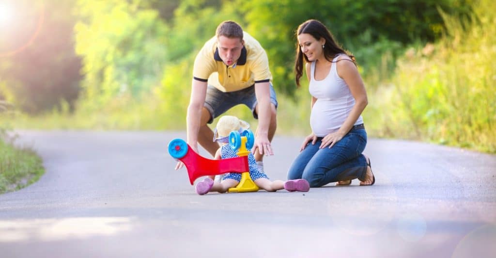 Parents assisting their toddler with a ride-on toy on an outdoor path.