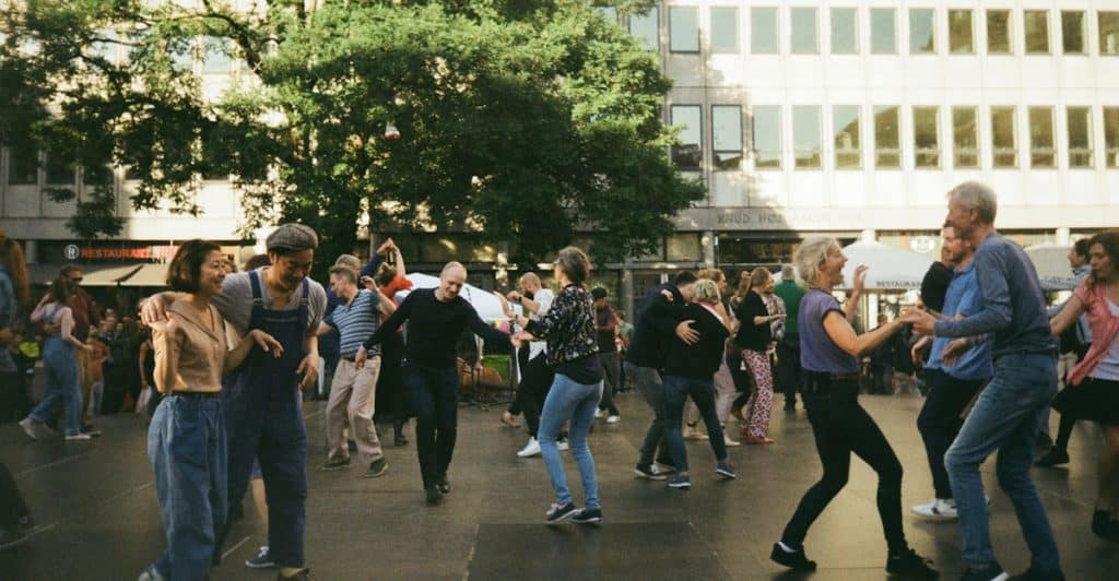 A large group of people dancing outdoors in a public square with trees and buildings in the background.
