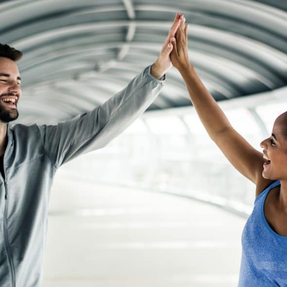 A man with a beard wearing a gray athletic jacket and a woman with dark skin and a blue tank top, giving each other a high-five in a bright, modern walkway with a curved ceiling.