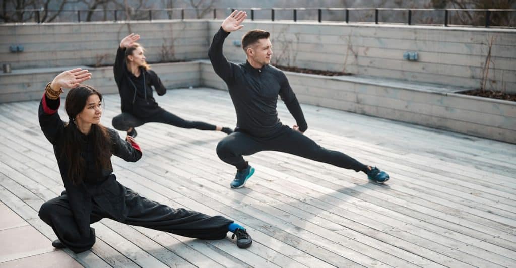 Three people dressed in dark, loose-fitting clothing practicing Tai Chi on an outdoor wooden platform.