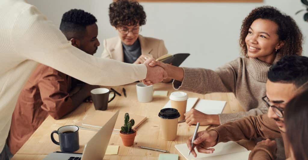 Two people shaking hands across a table in a meeting with other people present.