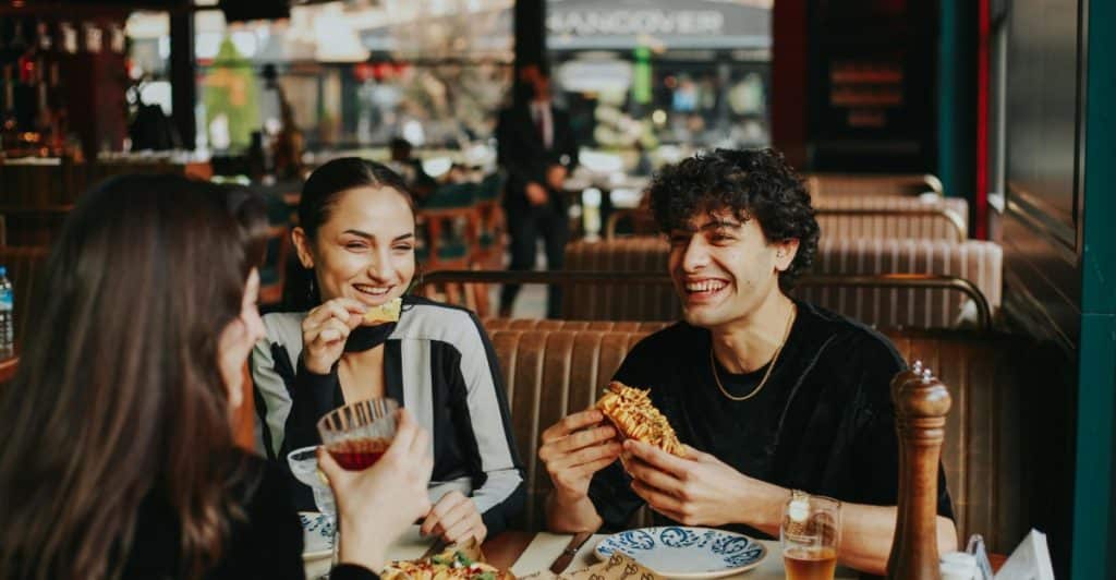 Three people eating and talking at a restaurant table with various dishes.