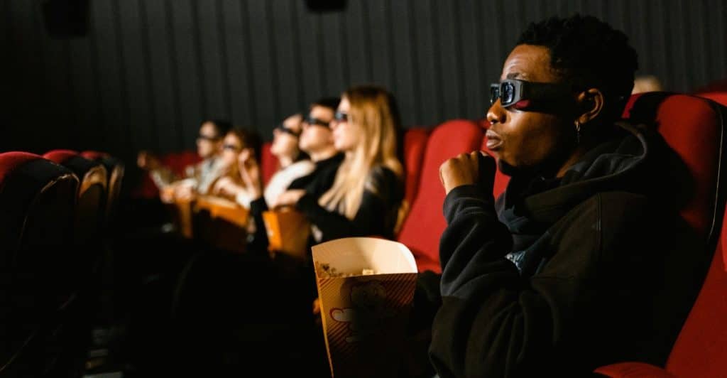 People in a dark movie theater wearing 3D glasses and watching a screen, with one person holding popcorn in the foreground.