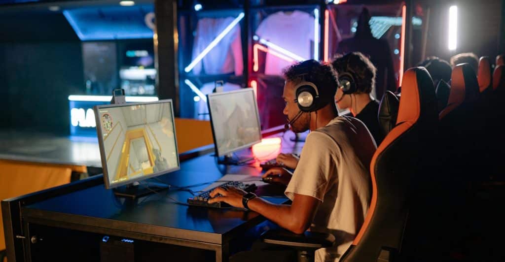 Two people playing first-person shooter games on computers in a gaming setup with headphones on.