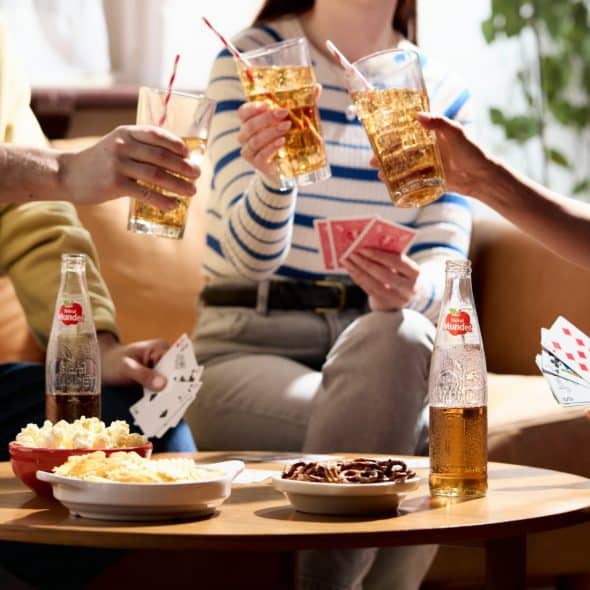 Four people gathered around a low round table, holding cards and drinks, with snacks on the table.