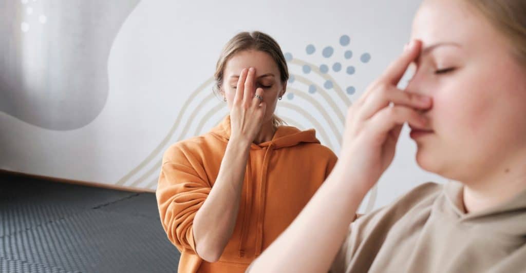 Two people, one in an orange hoodie and the other in a beige hoodie, practicing a breathing exercise while touching their faces.