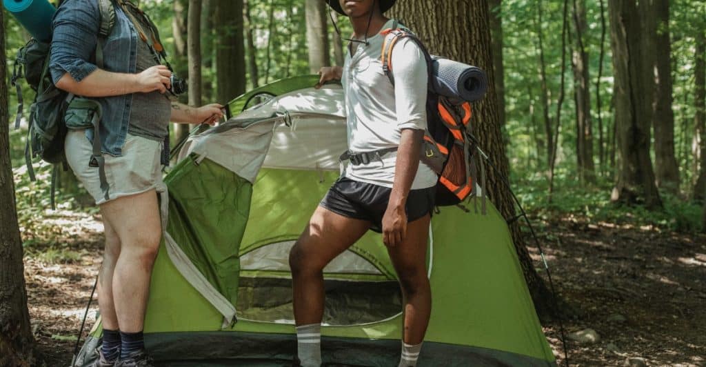 Two people with a set up a green tent in a forest, carrying hiking gear.