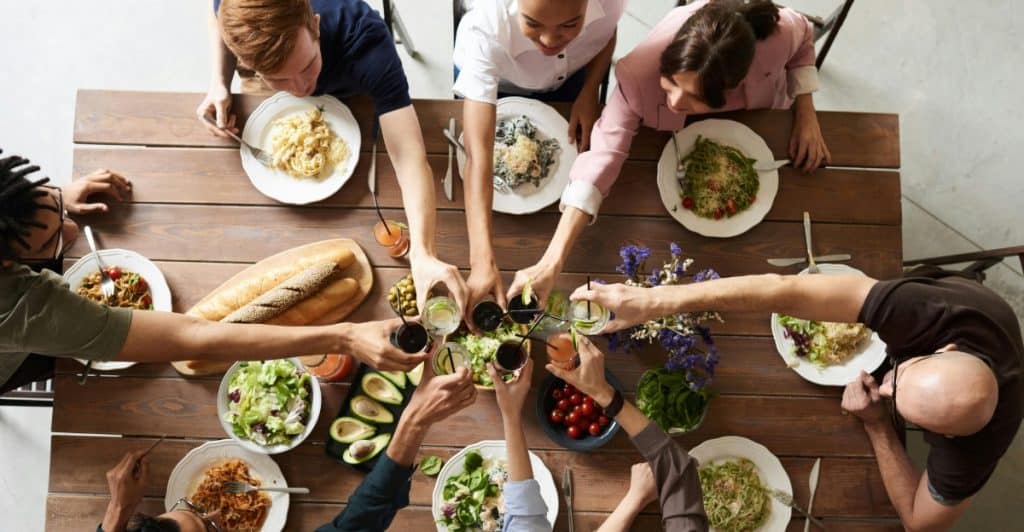 Overhead view of a group of people are sitting around a wooden table filled with food, raising their glasses in a toast.