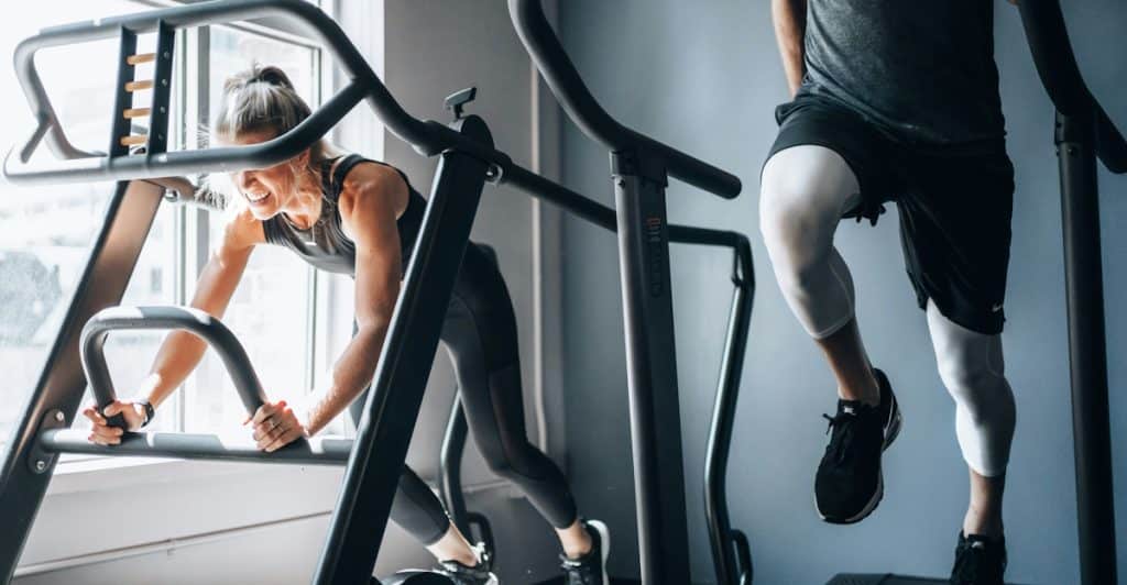 Two people on self-powered treadmills in a gym, with one person walking and the other using the machine.