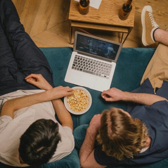 Two people relaxing on a teal couch, looking at a laptop and sharing a bowl of popcorn, with two bottles on a small wooden table behind the laptop.