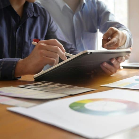 Two people are looking at color palettes and a tablet on a wooden table, with one person holding a pen.