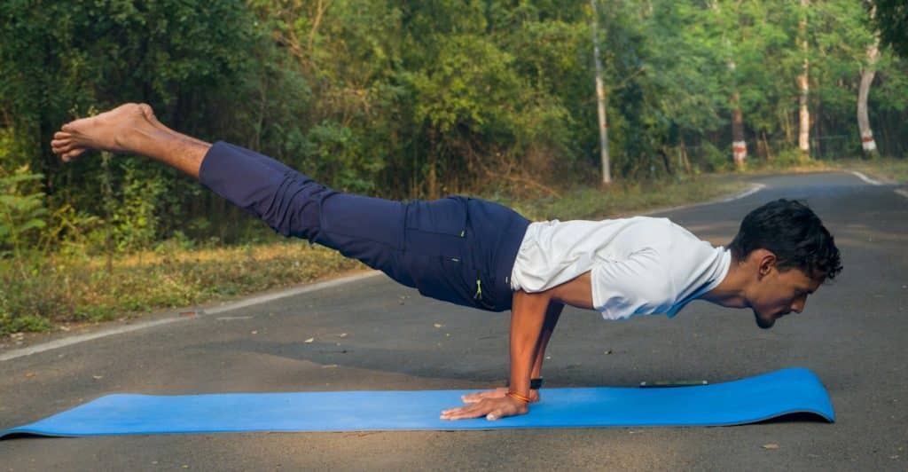 A person performing a balance exercise on a blue yoga mat on an outdoor paved surface, with a natural background of trees.