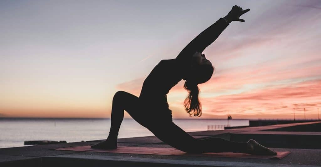 A silhouetted figure performing a yoga pose outdoors on a mat with a body of water and a colorful sunset in the background.