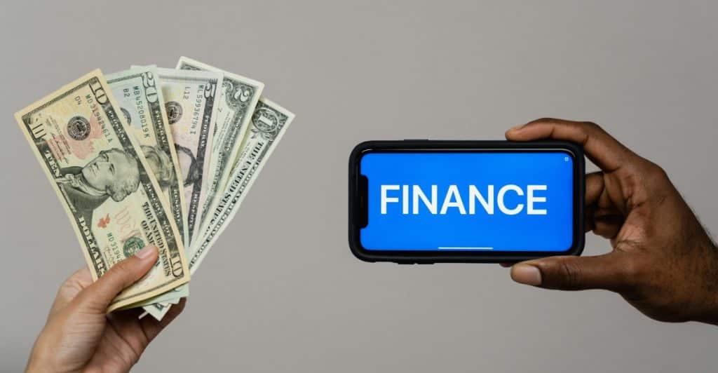 A person is holding a stack of cash in one hand and a phone with the word "FINANCE" on the screen in the other hand.