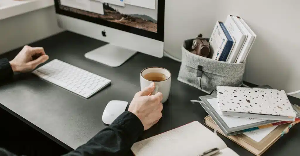 A person with a cup is writing in a journal at a desk with a computer.