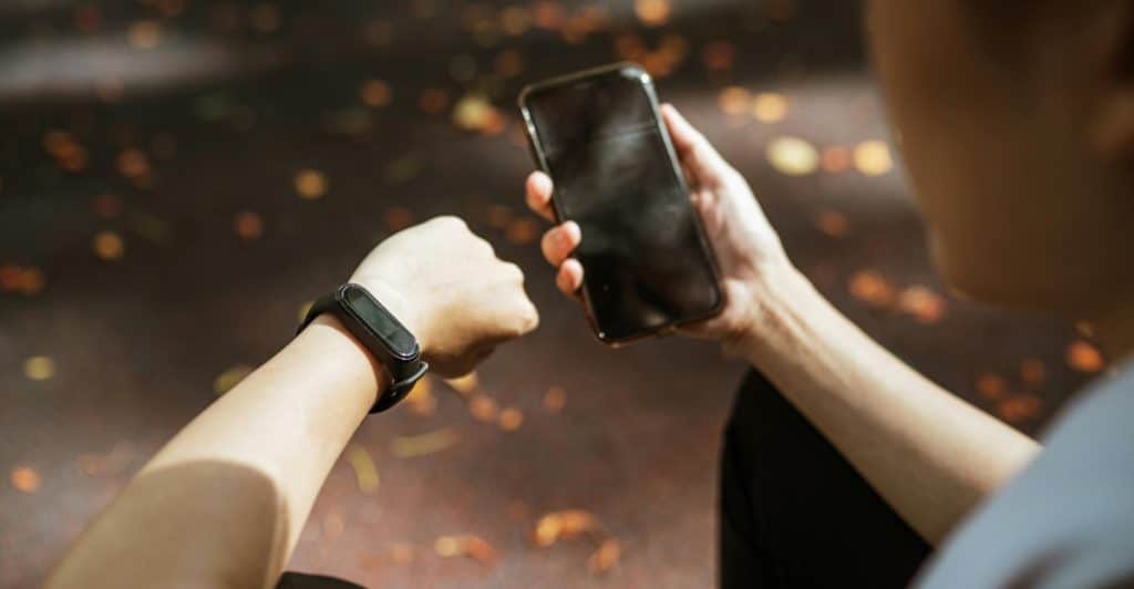 A close-up of a person's arm wearing a black fitness tracker watch, looking at their phone which is held in their other hand.
