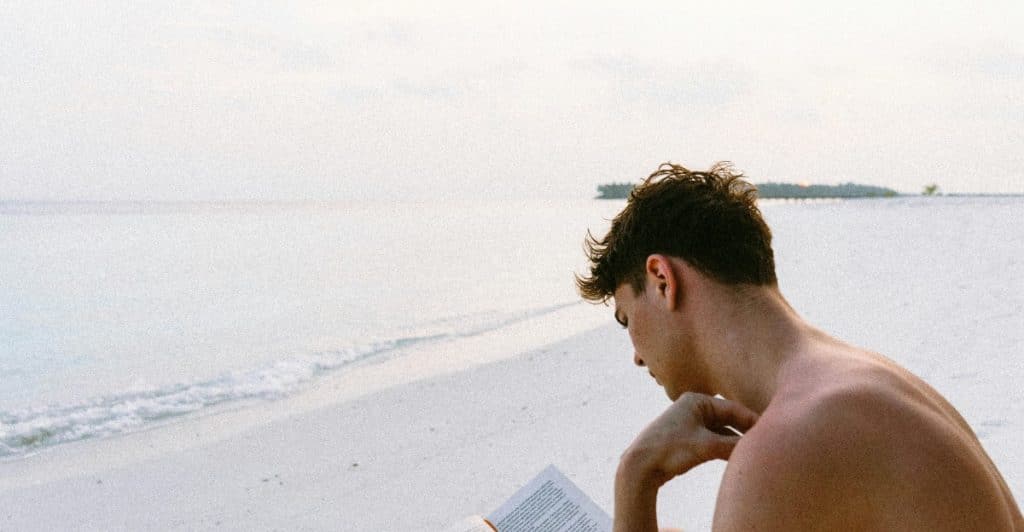 A person is seated on a light-colored beach, reading a book with the ocean and a distant shoreline in the background.