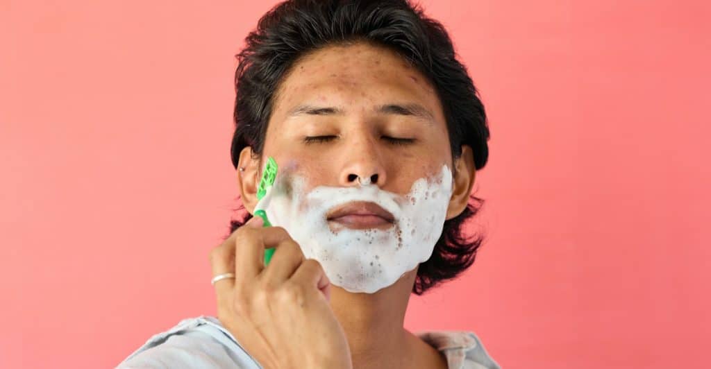 A person with shaving cream on their face, holding a razor against a pink background.