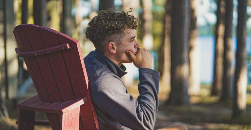 A person sitting in a red Adirondack chair outside, with a thoughtful expression, in a natural setting.