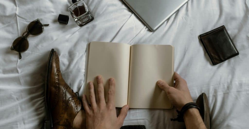 Person sitting on a bed with a notebook, laptop, sunglasses, and a fragrance bottle scattered around.