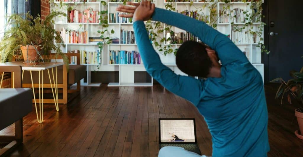 A person in front of a laptop stretching with their arms raised and clasped above their head on a yoga mat in a room with a bookshelf filled with books and plants in the background.