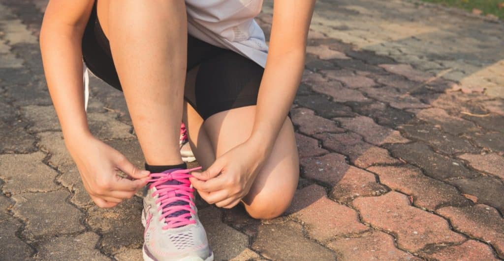Close-up of a person's hands tying the laces of a pink and gray running shoe on a paved surface.