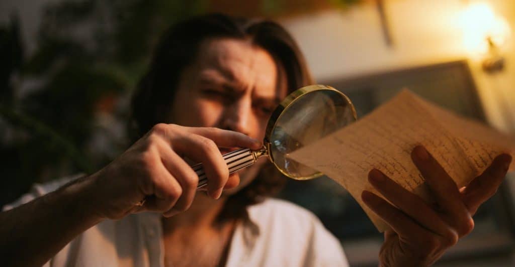 A person with light brown hair is shown in a close-up, holding a large magnifying glass to examine text on a piece of aged paper, with a focused expression.