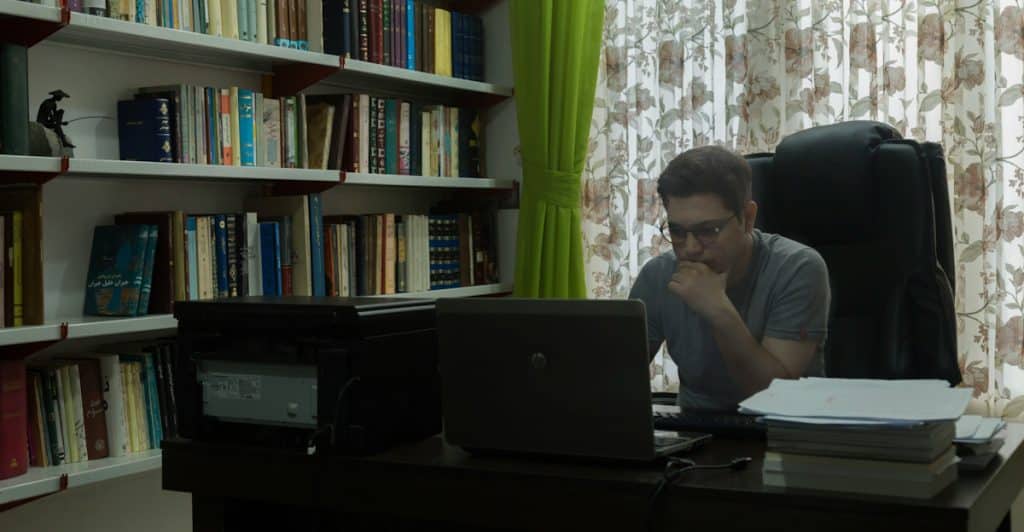 A person wearing glasses sitting at a desk with a laptop and papers, in a room with bookshelves and curtains.