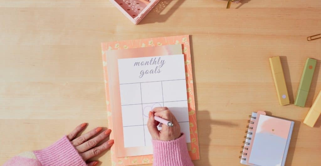 Overhead shot of a person's hands writing "monthly goals" on a structured planner surrounded by stationery items on a wooden desk.