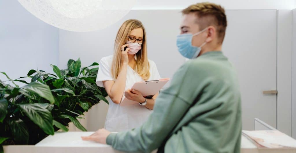 A person wearing a green jacket and a face mask standing at a reception desk talking to a healthcare worker wearing a white coat and face mask.