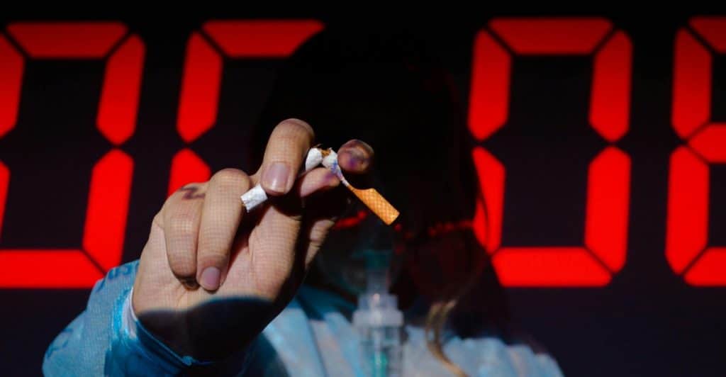 A close-up of a hand holding a lit cigarette, with red digital numbers in the background.