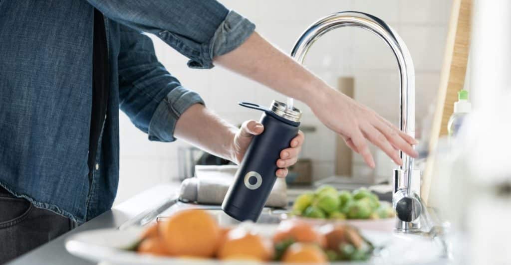 A person wearing a denim shirt fills a dark blue water bottle at a kitchen sink.