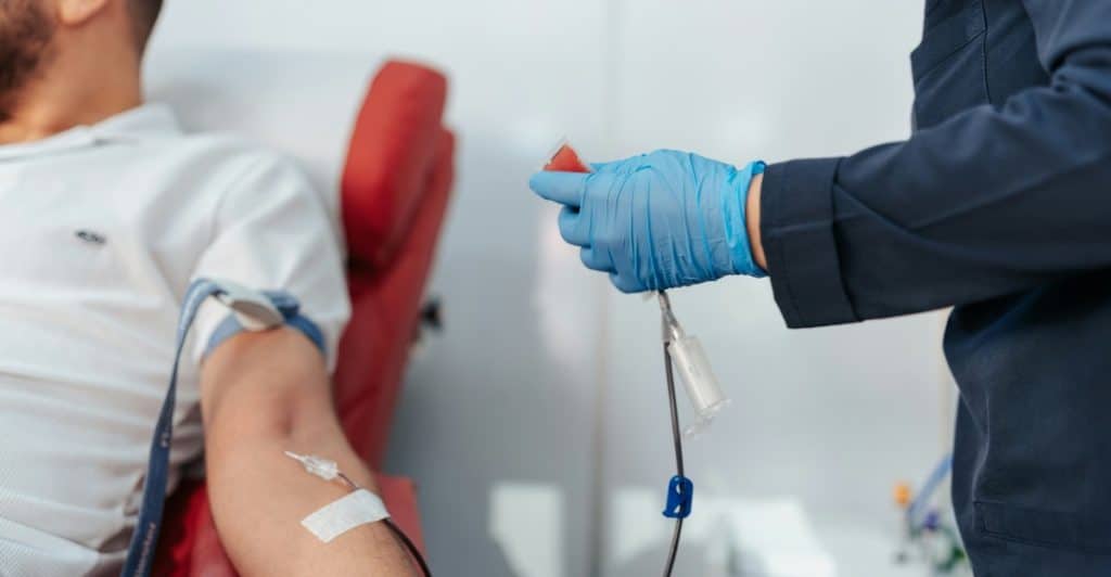 A close-up of a person's arm with a needle inserted for blood draw, and a gloved hand holding a test tube.
