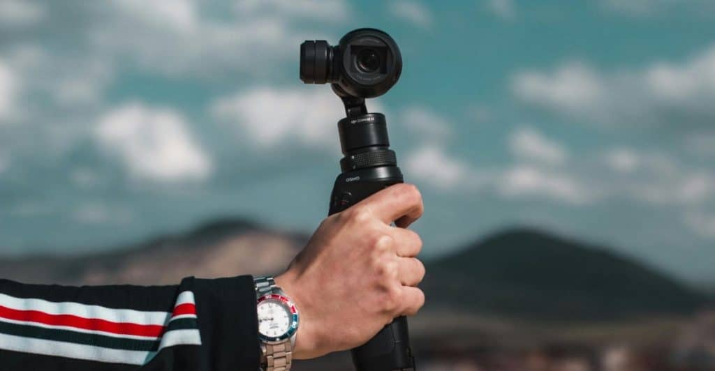 A person's hand wearing a watch and a black jacket with red and white stripes holding a DJI Osmo Pocket 3, with a blurred outdoor background.