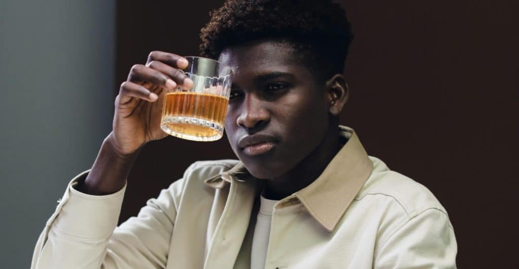 A person with dark skin and dark curly hair, wearing a light-colored jacket, holding a glass with an amber-colored liquid.
