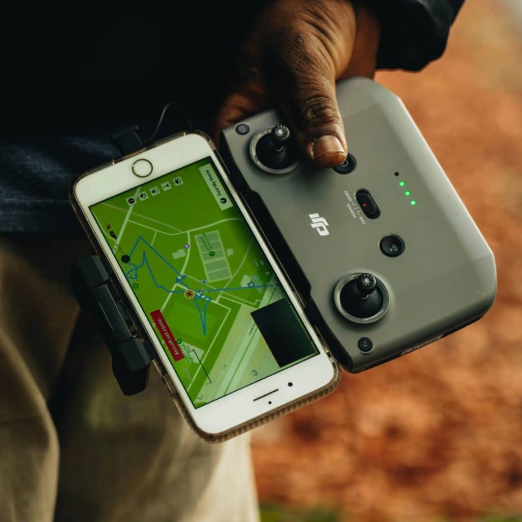 A close-up shot of a person's hand holding a white smartphone displaying a map and a gray drone controller, with a blurred outdoor background.