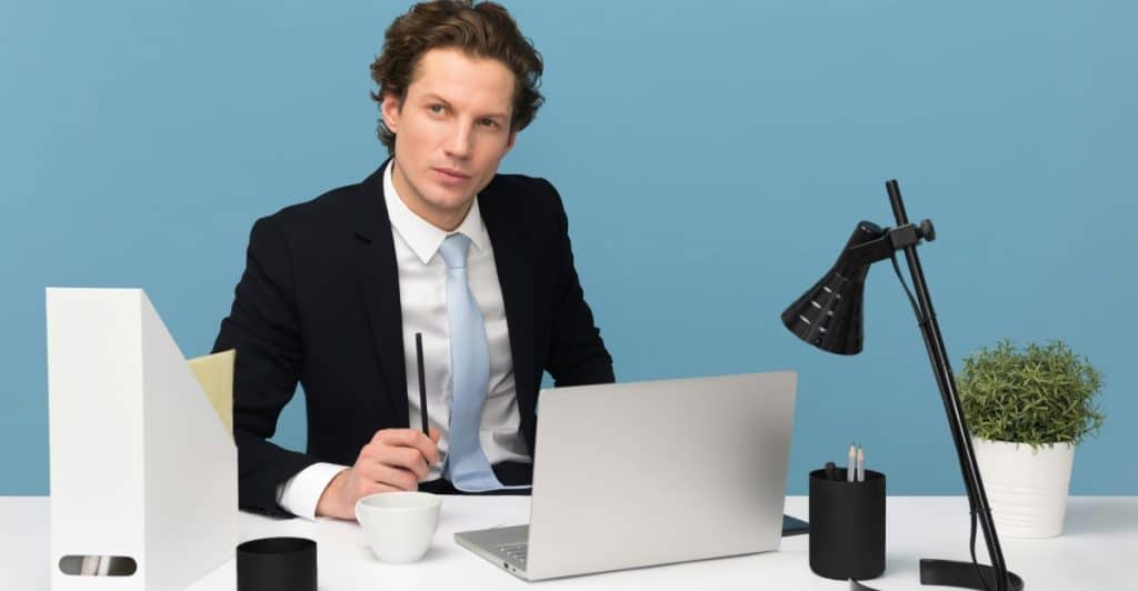 A person in business attire sits at a desk with a laptop, pens, and office supplies against a blue background.
