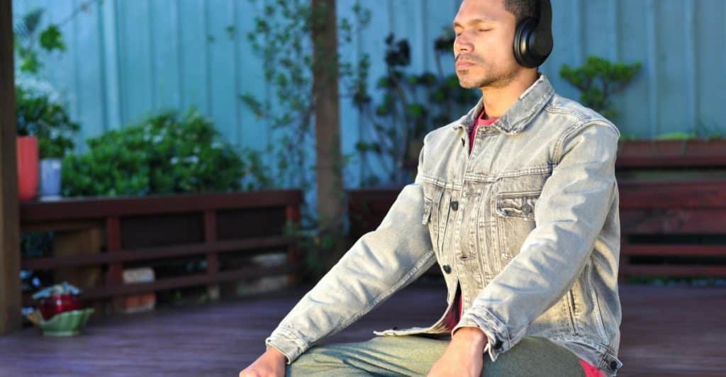 A person with dark hair wearing a denim jacket and headphones meditating in a seated position outdoors.