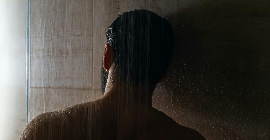 A person with dark hair standing with their back to the viewer in a shower with water running over them.