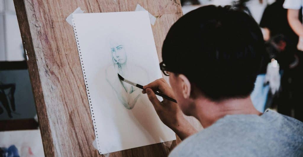 A person sketching a portrait on a canvas with a brush in an art studio setting.