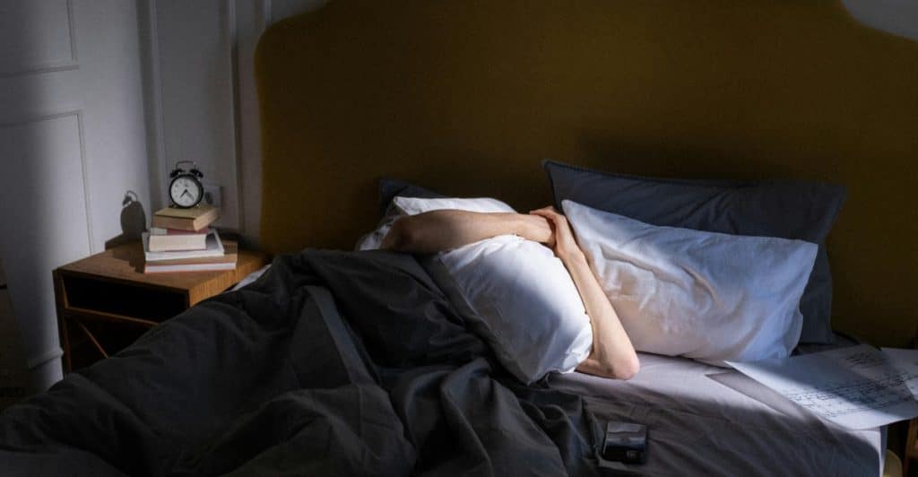 A person lying on a bed holding a pillow against his face in a dimly lit room with an alarm clock and papers on the bedside table.