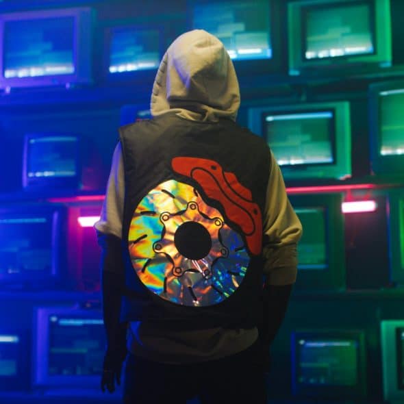 A person wearing a light-hooded sweatshirt and a dark vest with a large, colorful reflective design on the back is standing facing away from the viewer in front of a wall of stacked old-fashioned computer monitors emitting blue and green light.