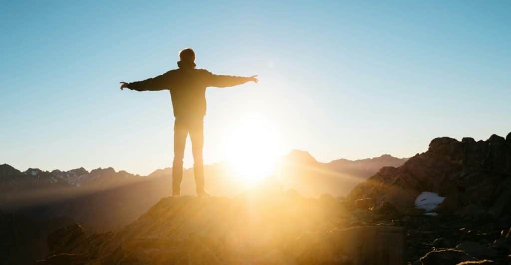 A person standing on a mountaintop with outstretched arms, facing a sunrise.