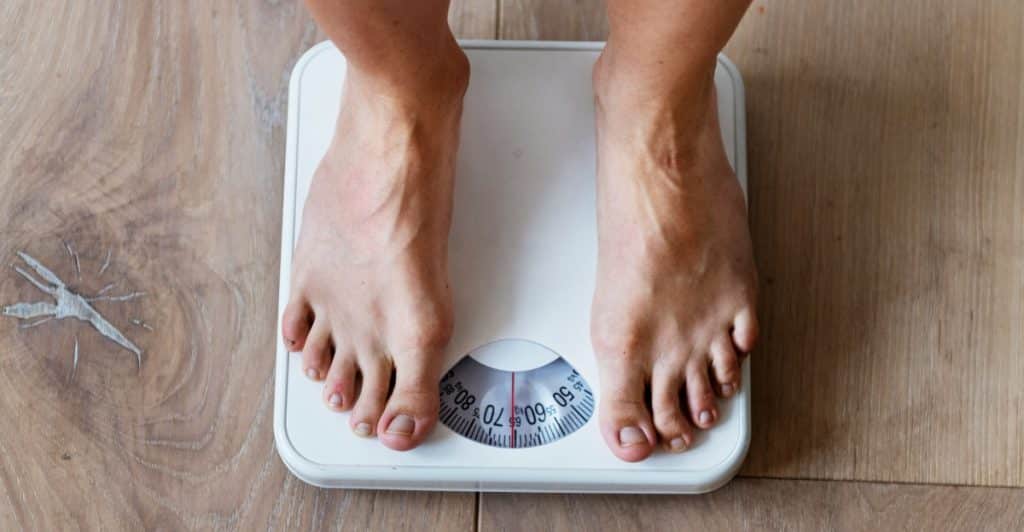 A close-up of a person's feet standing on a white weighing scale on a wooden floor.