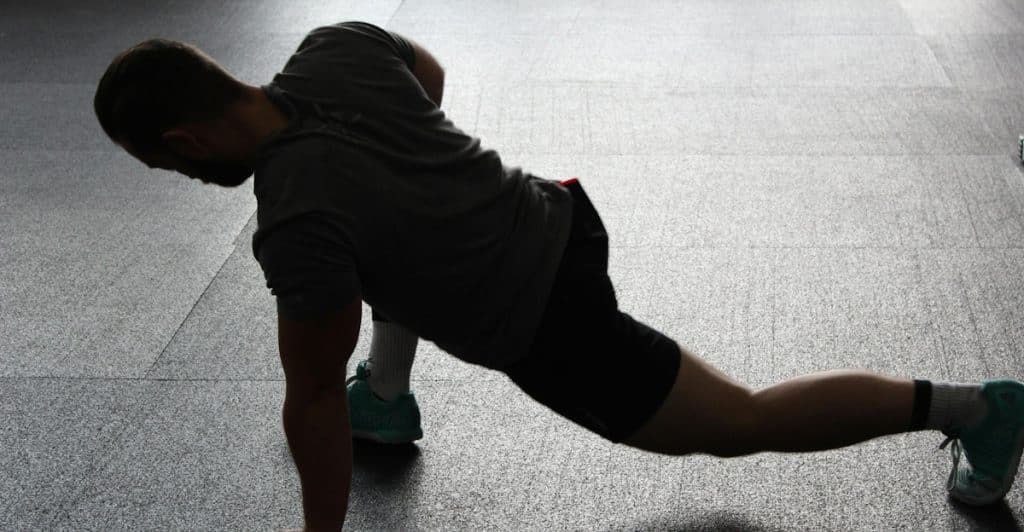 A person in athletic wear performing a stretching exercise on a gym floor, silhouetted against the light.