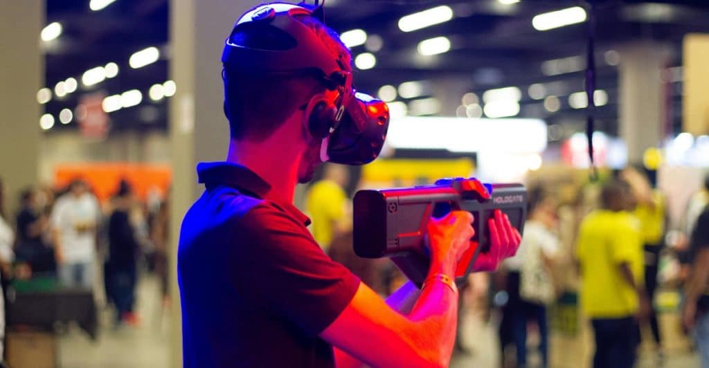 A person wearing a virtual reality headset and holding a gun-shaped controller in a crowded space.