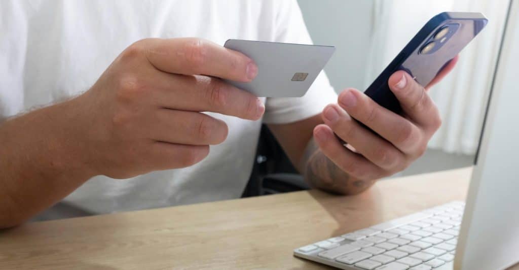 A person holding a credit card and using a smartphone while sitting at a desk with a computer.