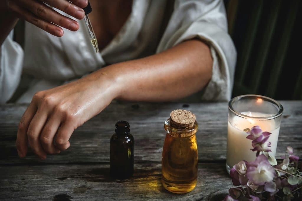 A person in a white robe using a dropper to apply essential oil to their arm, with bottles and a candle nearby.
