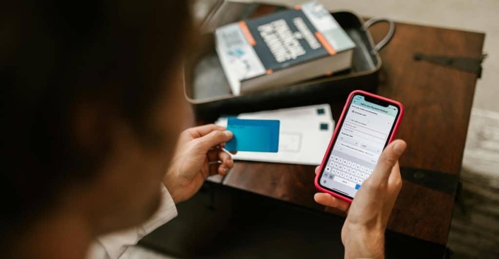 A person is using a smartphone with a credit card and a book on financial planning nearby.