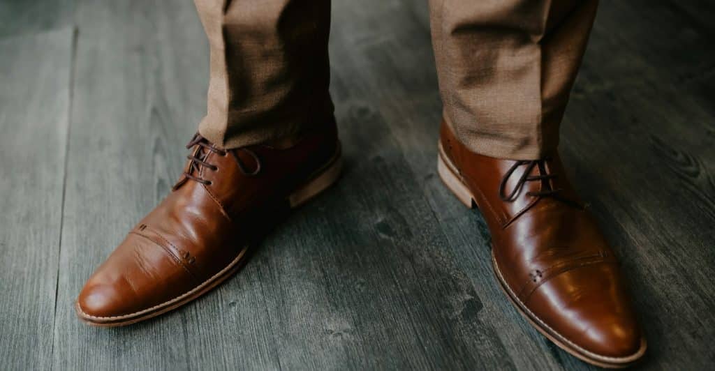 A person standing on a wooden floor wearing brown dress shoes.